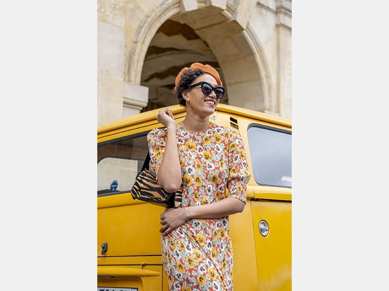 Femme souriante en robe fleurie et béret orange, posant devant une voiture jaune stationnée sous une arche.