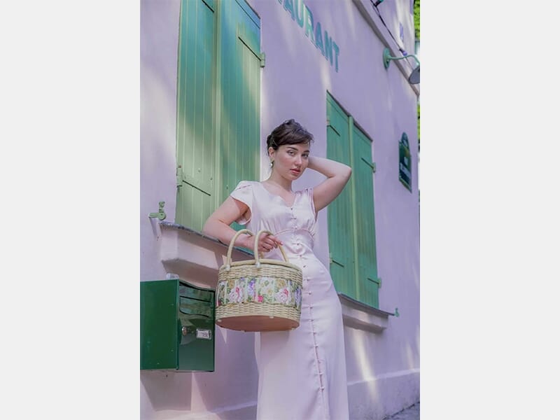 Femme en robe claire tenant un panier en osier décoré, posée devant un mur rose aux volets verts.