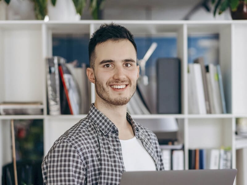 Jeune homme souriant en chemise à carreaux, tenant un ordinateur portable devant une étagère.
