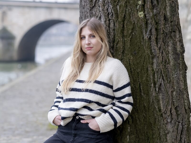 Femme en pull rayé blanc et bleu marine, appuyée contre un arbre au bord de la Seine, regard calme.