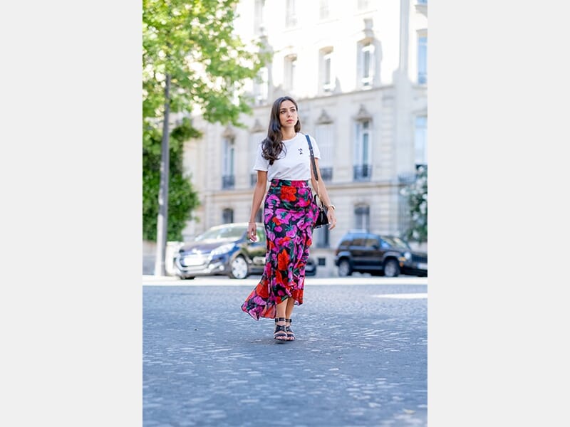 Femme marchant sur une rue pavée, portant un t-shirt blanc et une jupe longue à motifs floraux colorés.