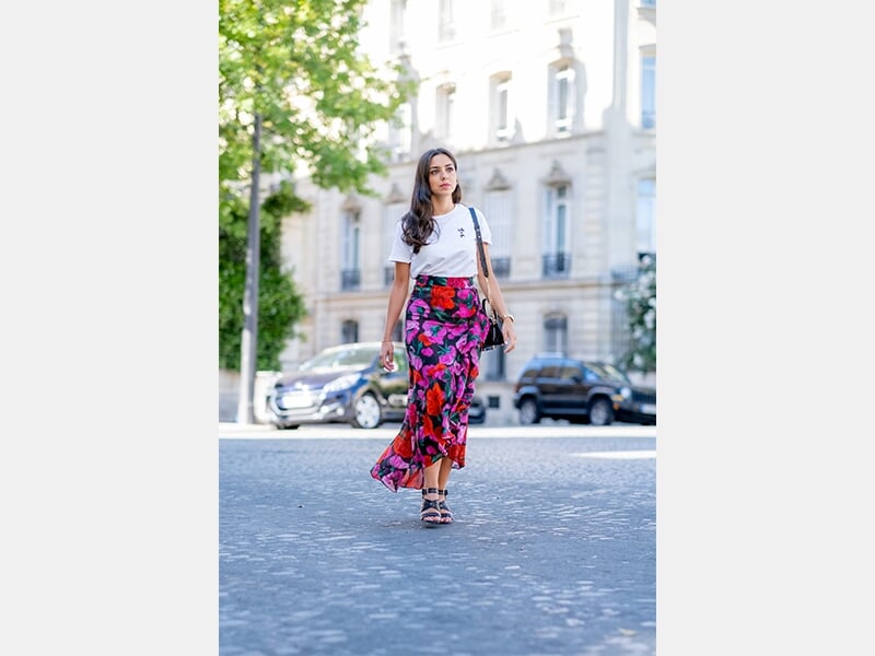 Femme marchant sur une rue pavée, portant un t-shirt blanc et une jupe longue à motifs floraux colorés.