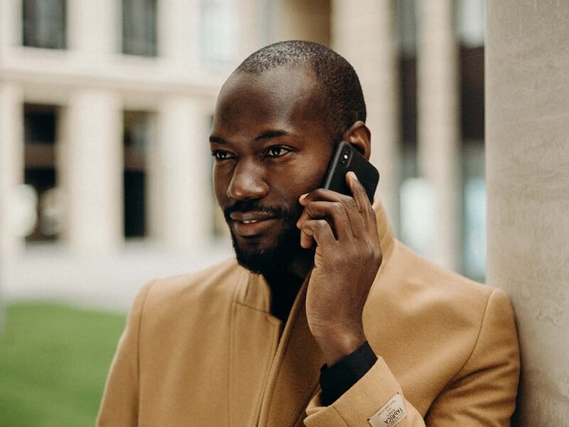 Homme souriant en manteau beige, parlant au téléphone portable, adossé à une colonne en ville.