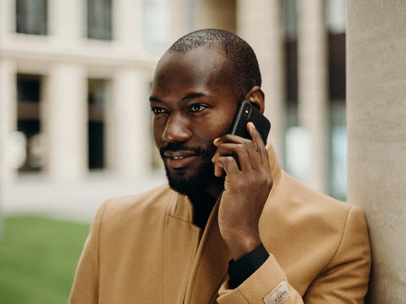 Homme souriant en manteau beige, parlant au téléphone portable, adossé à une colonne en ville.