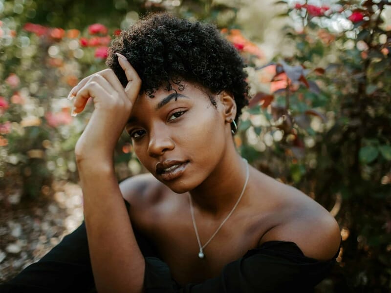 Femme assise dans un jardin fleuri, regard intense, main posée sur le front, robe noire tombant des épaules.
