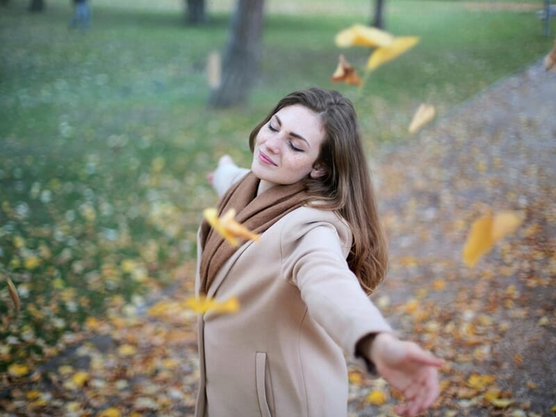 Femme souriante en manteau beige, bras ouverts sous des feuilles jaunes tombantes dans un parc.