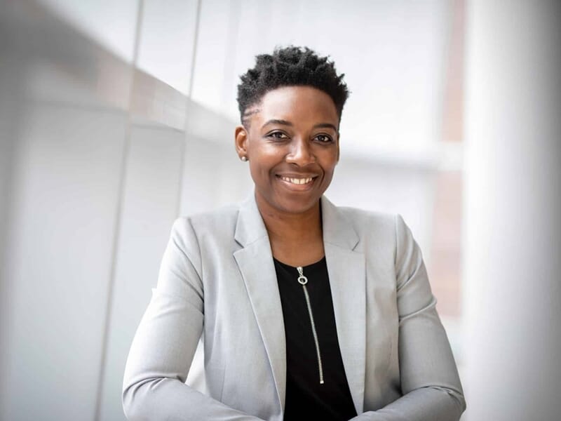 Femme souriante en blazer gris et haut noir, posée contre une paroi vitrée blanche en intérieur.