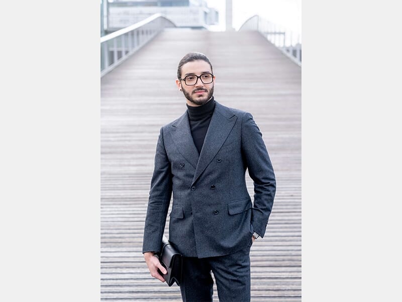 Homme barbu en costume croisé gris foncé et col roulé noir, debout sur un large escalier en bois.