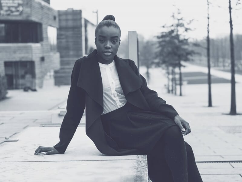 Femme assise sur un banc urbain, portant un manteau sombre et chemise blanche, regard sérieux, photo en noir et blanc.
