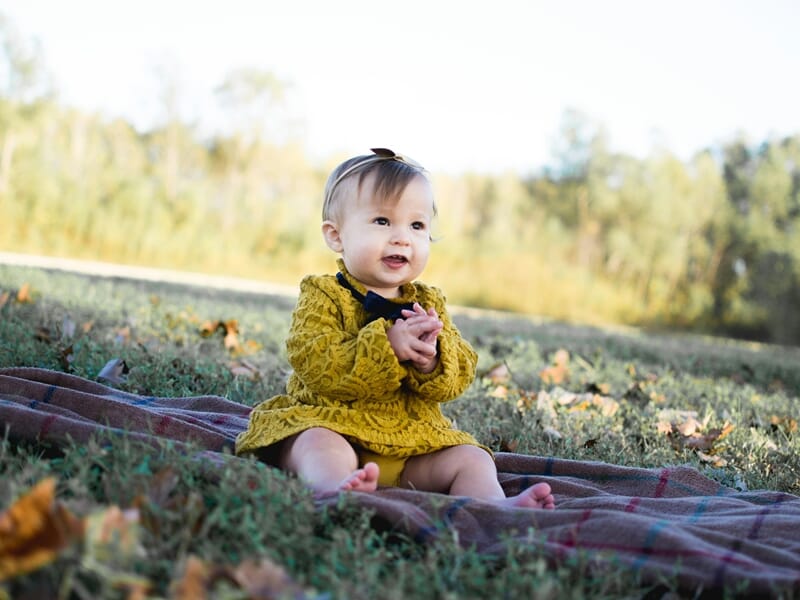 Bébé assise sur une couverture à carreaux dans l’herbe, portant une robe en dentelle jaune moutarde avec un nœud noir, mains jointes et expression joyeuse, fond de forêt floue.