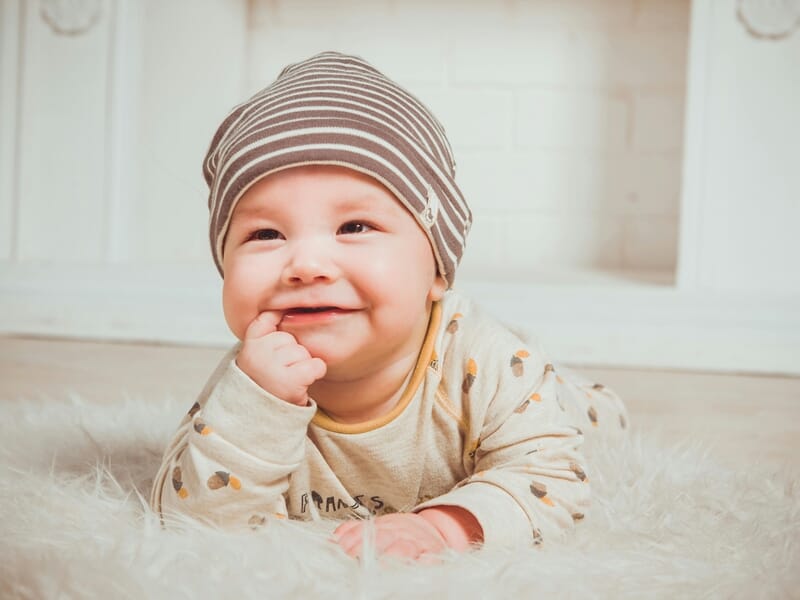 Bébé allongé sur un tapis en fausse fourrure blanche, portant un bonnet rayé marron et blanc et un pyjama beige à motifs, sourire aux lèvres et doigt dans la bouche.