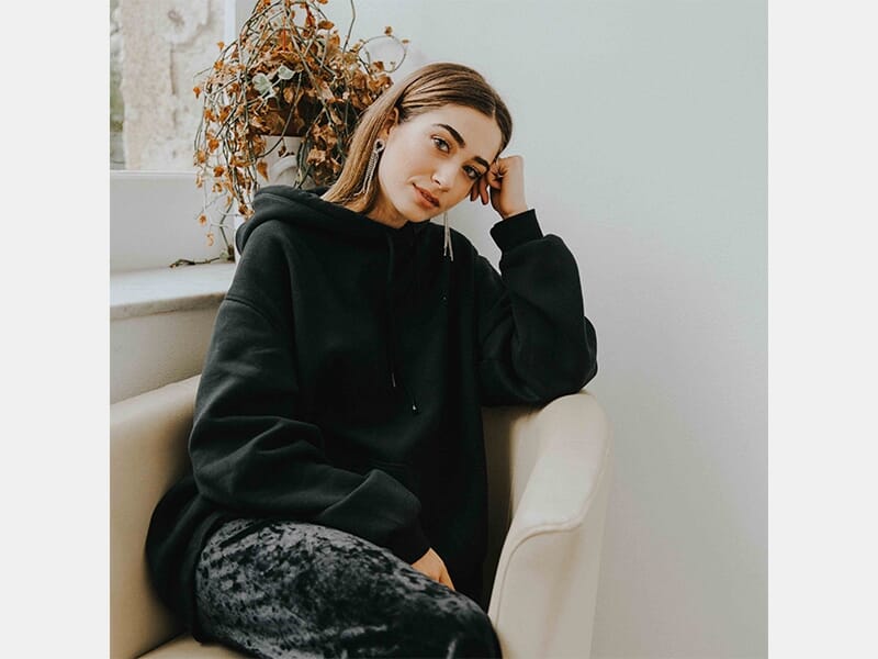 Femme assise dans un fauteuil près d'une fenêtre, portant un sweat noir à capuche, regard calme et pensif.