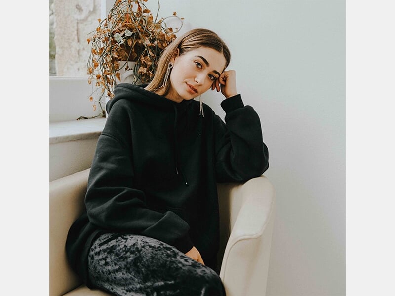 Femme assise dans un fauteuil près d'une fenêtre, portant un sweat noir à capuche, regard calme et pensif.