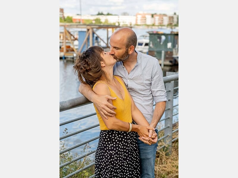 Couple s'embrassant tendrement devant une rambarde au bord d'un port, avec des bateaux et des immeubles en arrière-plan.