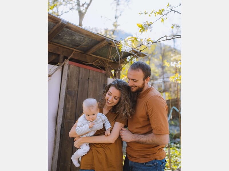 Jeune couple souriant avec leur bébé dans les bras, debout à l'extérieur près d'une cabane en bois, par une journée ensoleillée.