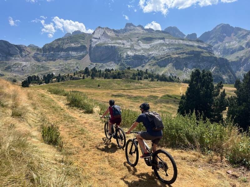 Deux cyclistes en VTT sur une piste herbeuse, avec les montagnes et le ciel bleu en fond.