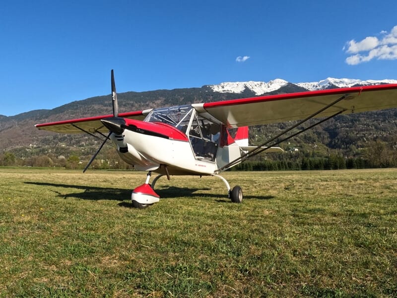 Petit avion blanc et rouge posé sur un champ herbeux, avec en arrière-plan des montagnes partiellement enneigées sous un ciel bleu.