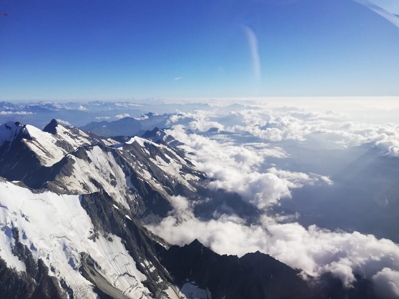 Paysage de montagne enneigée vu du ciel, avec des sommets émergeant d’une mer de nuages sous un ciel bleu pâle.