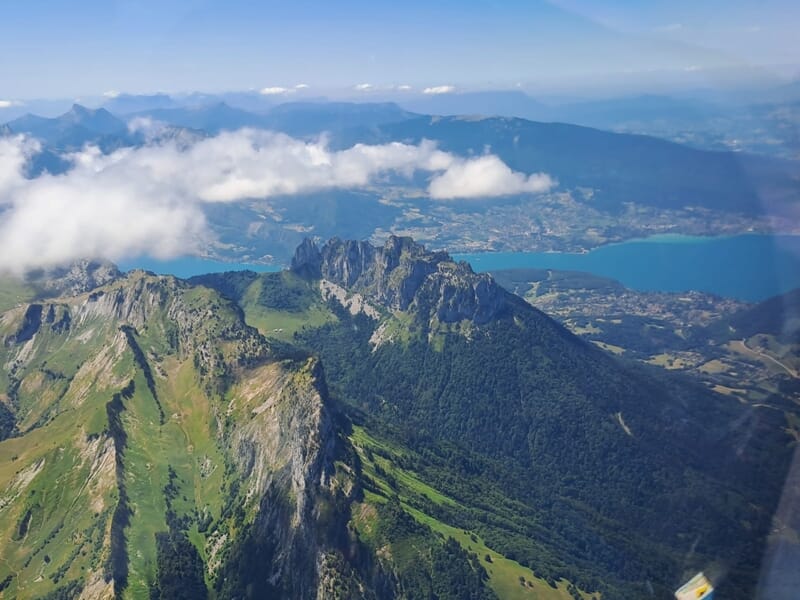 Chaîne de montagnes verdoyantes surplombant un lac turquoise, vue depuis les airs avec des nuages bas.