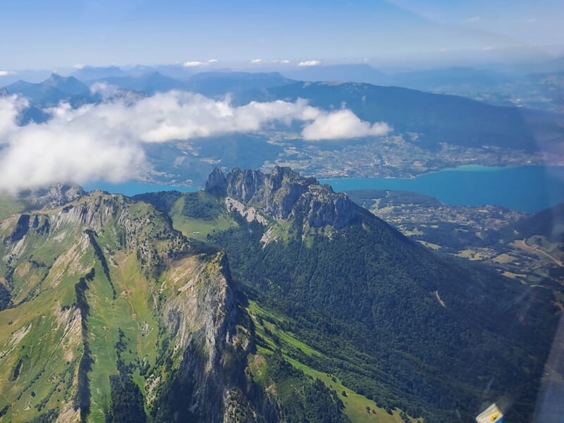 Chaîne de montagnes verdoyantes surplombant un lac turquoise, vue depuis les airs avec des nuages bas.