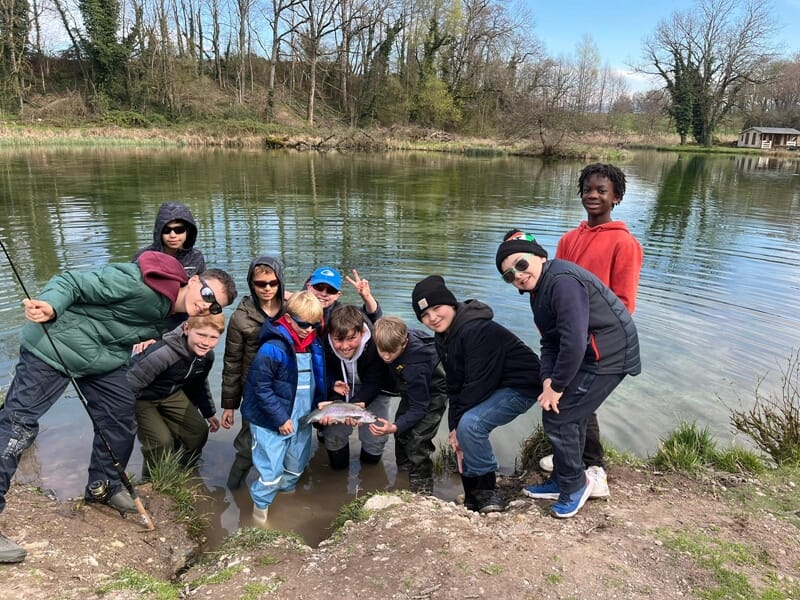 Groupe d’enfants au bord d’un étang, rassemblés autour d’un adulte tenant une truite lors d’un atelier pêche.
