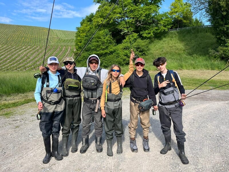 Six adolescents en tenue de pêche à la mouche posant avec leurs cannes devant un champ et des vignes.