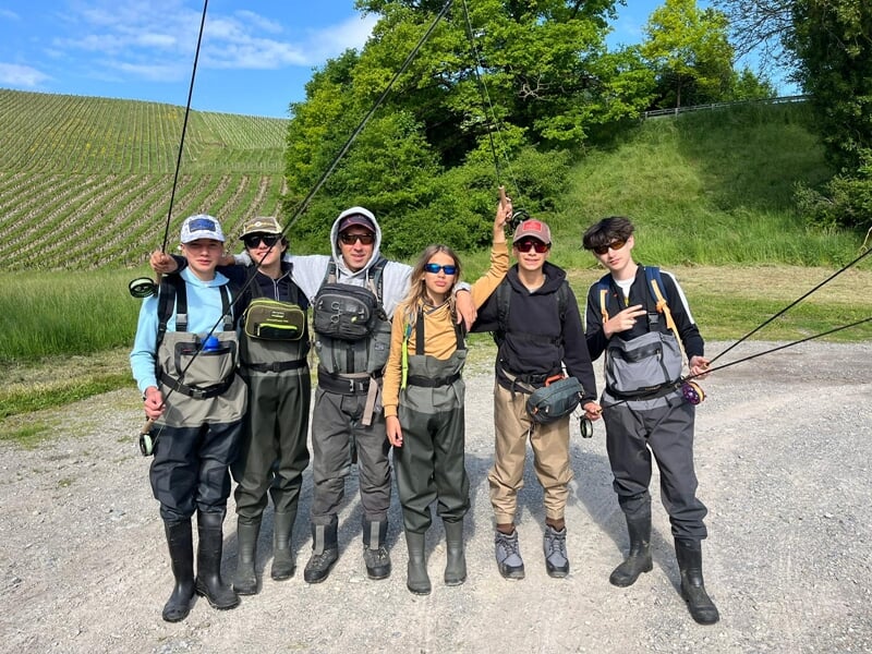 Six adolescents en tenue de pêche à la mouche posant avec leurs cannes devant un champ et des vignes.