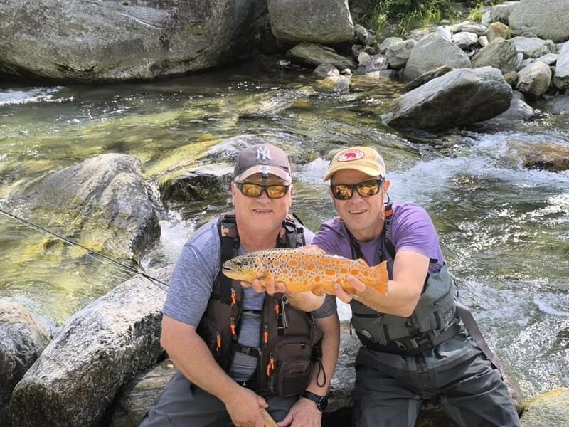 Deux hommes accroupis au bord d’un torrent tenant ensemble une truite fario orange aux points bien marqués.