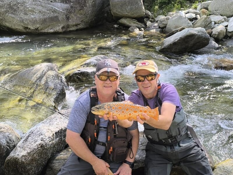 Deux hommes accroupis au bord d’un torrent tenant ensemble une truite fario orange aux points bien marqués.