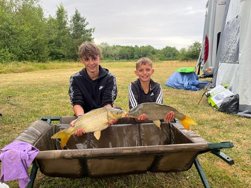Deux garçons souriants tenant chacun une carpe devant un tapis de réception, en plein air près d'une tente de camping.