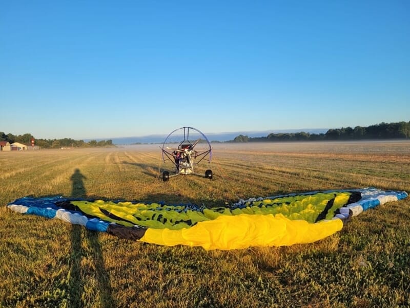 Paramoteur prêt à décoller sur une prairie au petit matin, avec la voile jaune et bleue déployée au sol