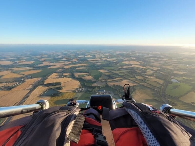 Vue en vol depuis un paramoteur, avec un sac à dos au premier plan et un paysage de champs à perte de vue sous un ciel dégagé