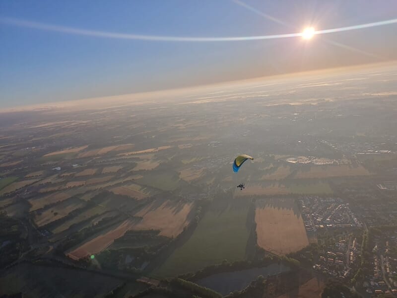 Vue aérienne d’un paramoteur en vol au lever du soleil, survolant une mosaïque de champs et de villages