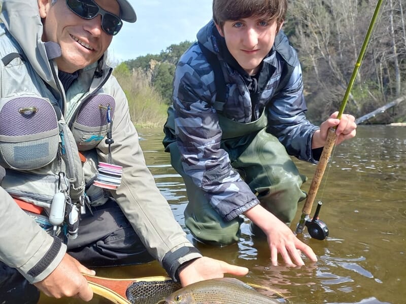 Père et fils en tenue de pêche montrant une prise dans une rivière, sourire et complicité