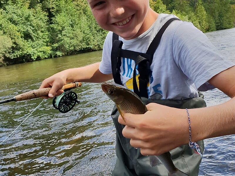 Jeune garçon souriant tenant un poisson fraîchement pêché en rivière avec une canne à mouche