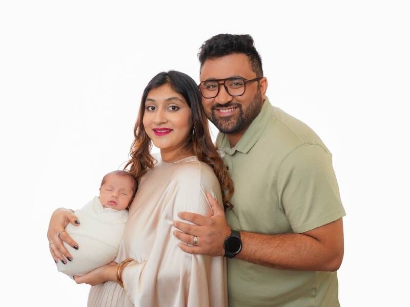 Portrait d'une famille souriante avec un bébé emmailloté, posant ensemble devant un fond blanc uni.