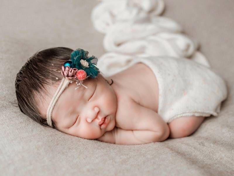 Nouveau-né endormi sur le côté, portant un bandeau fleuri, allongé sur un tissu beige doux.