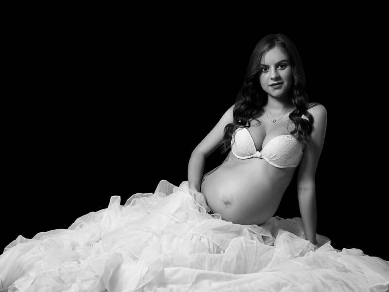 Une femme enceinte en lingerie blanche est assise sur un tissu blanc, regardant l'objectif avec douceur.