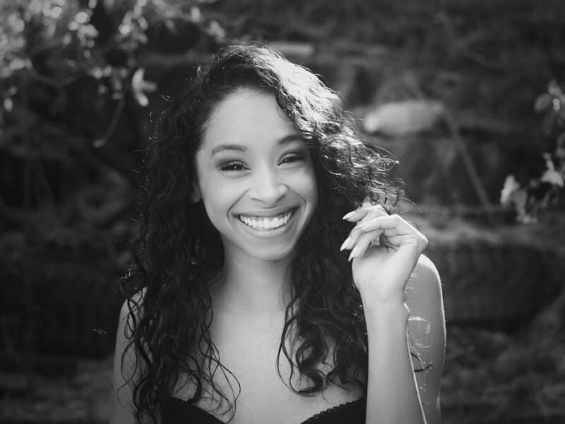 Portrait en noir et blanc d'une femme souriante aux cheveux bouclés, en extérieur avec un fond flou naturel.