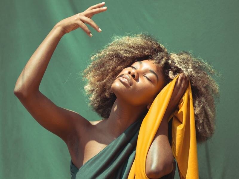 Femme aux cheveux afro posant avec un tissu jaune et vert, yeux fermés, sur fond vert uni, lumière naturelle.