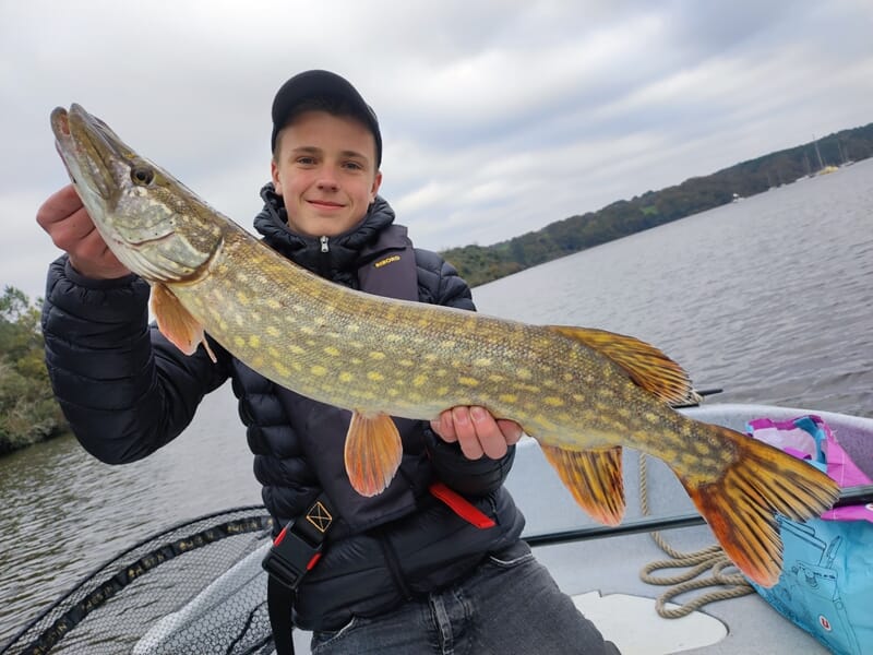Un adolescent en tenue de pêche tient un grand brochet tacheté, fraîchement sorti de l’eau, sur un bateau.