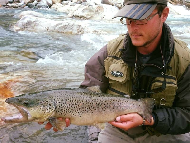 Un homme en tenue de pêche, au bord d'une rivière, présente une belle truite fario.