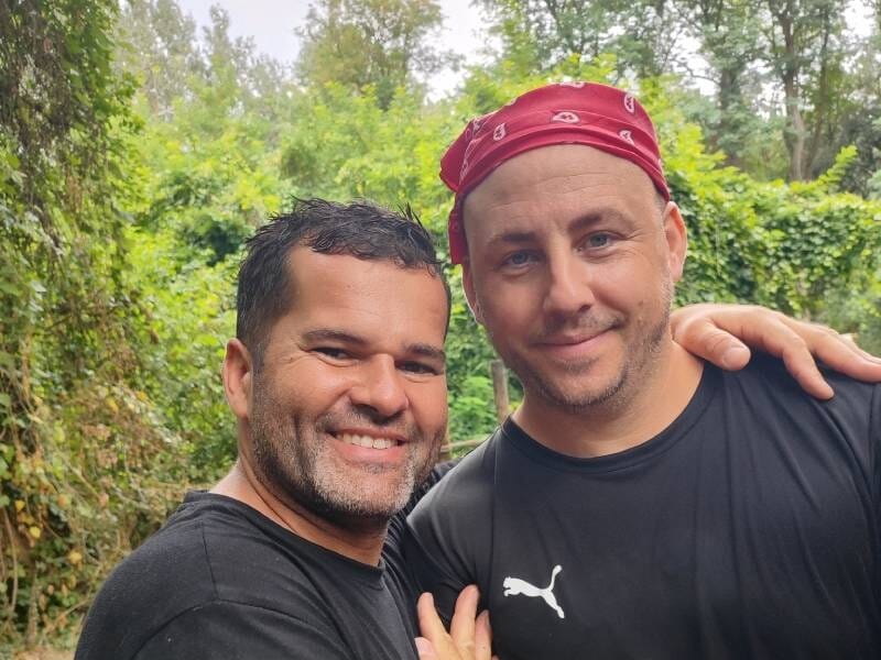 Deux hommes souriants, l'un a ses bras autour des épaules de l'autre, qui porte un bandana rouge.