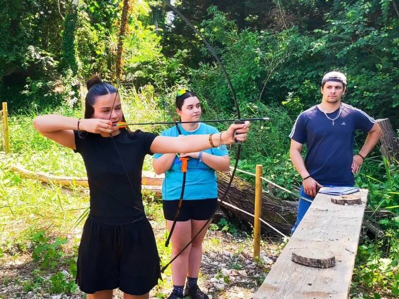 Une jeune femme en train de tirer à l'arc et deux personnes qui l'observent.