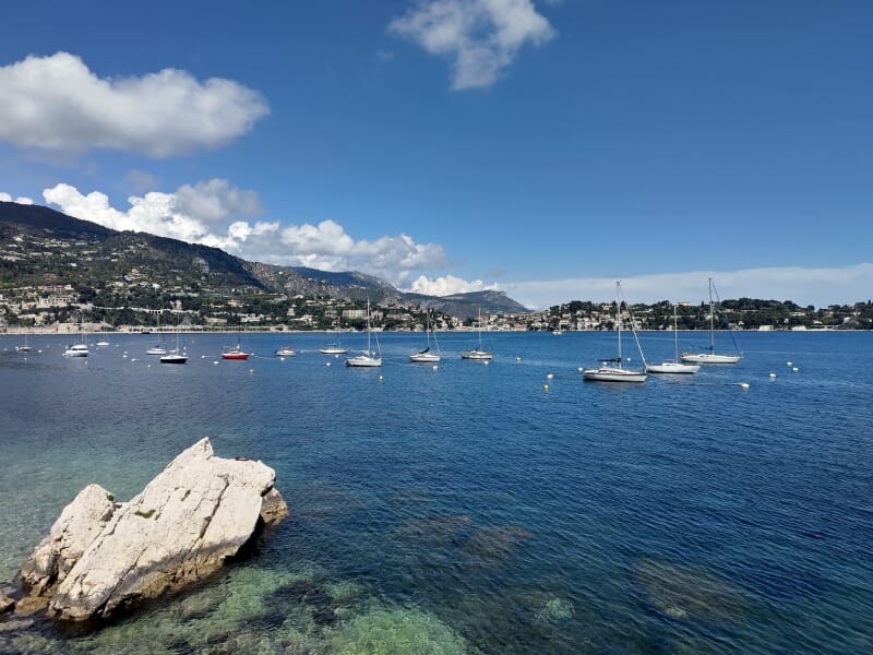 Vue panoramique sur une baie méditerranéenne avec des voiliers et collines en arrière-plan.