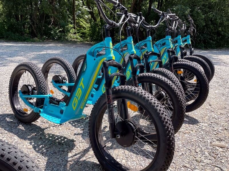 Plusieurs trottinettes électriques bleues alignées sur un sol en gravier sous un ciel ensoleillé.