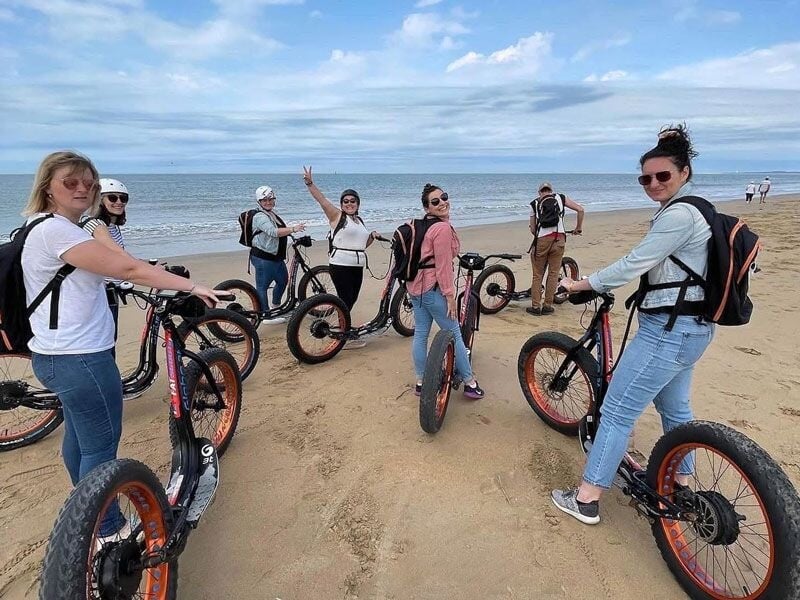 Groupe de personnes avec des trottinettes électriques sur une plage, posant et souriant vers la caméra.