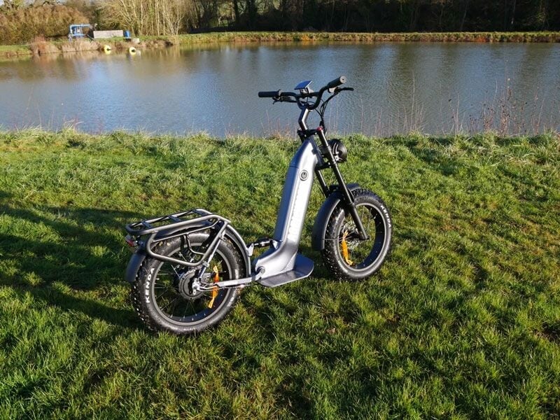Trottinette électrique grise garée sur une pelouse verte près d'un plan d'eau sous un ciel partiellement nuageux.