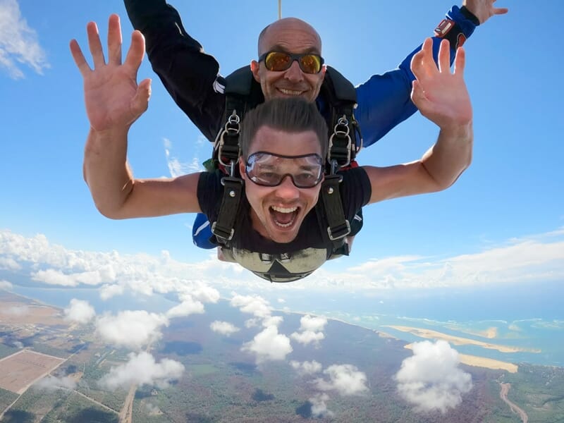 Tandem de parachutisme en plein vol, les deux hommes souriants face à la caméra avec un paysage côtier et nuageux en arrière-plan.