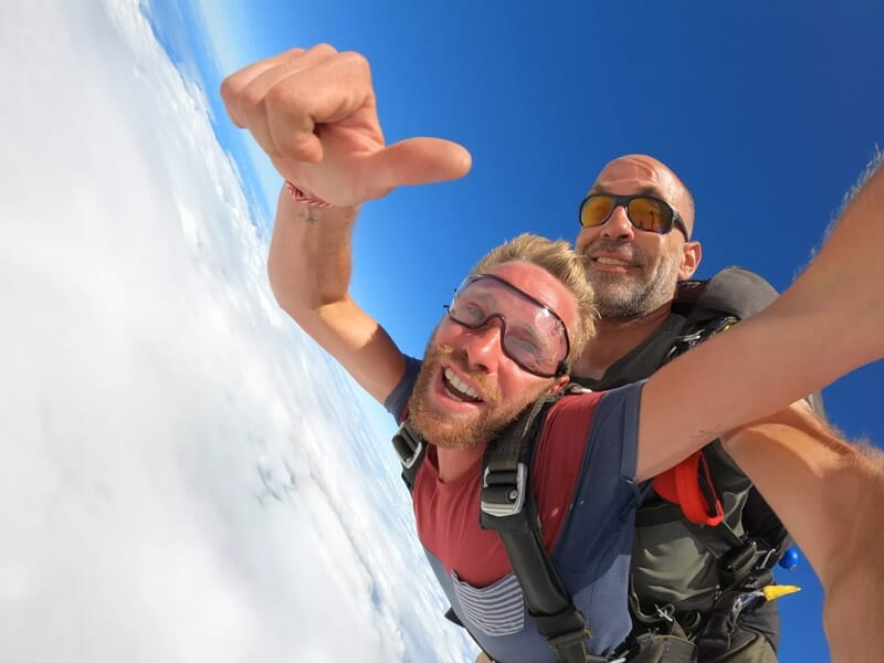 Deux hommes souriants en chute libre au-dessus d'une mer de nuages blancs sous un ciel bleu éclatant.